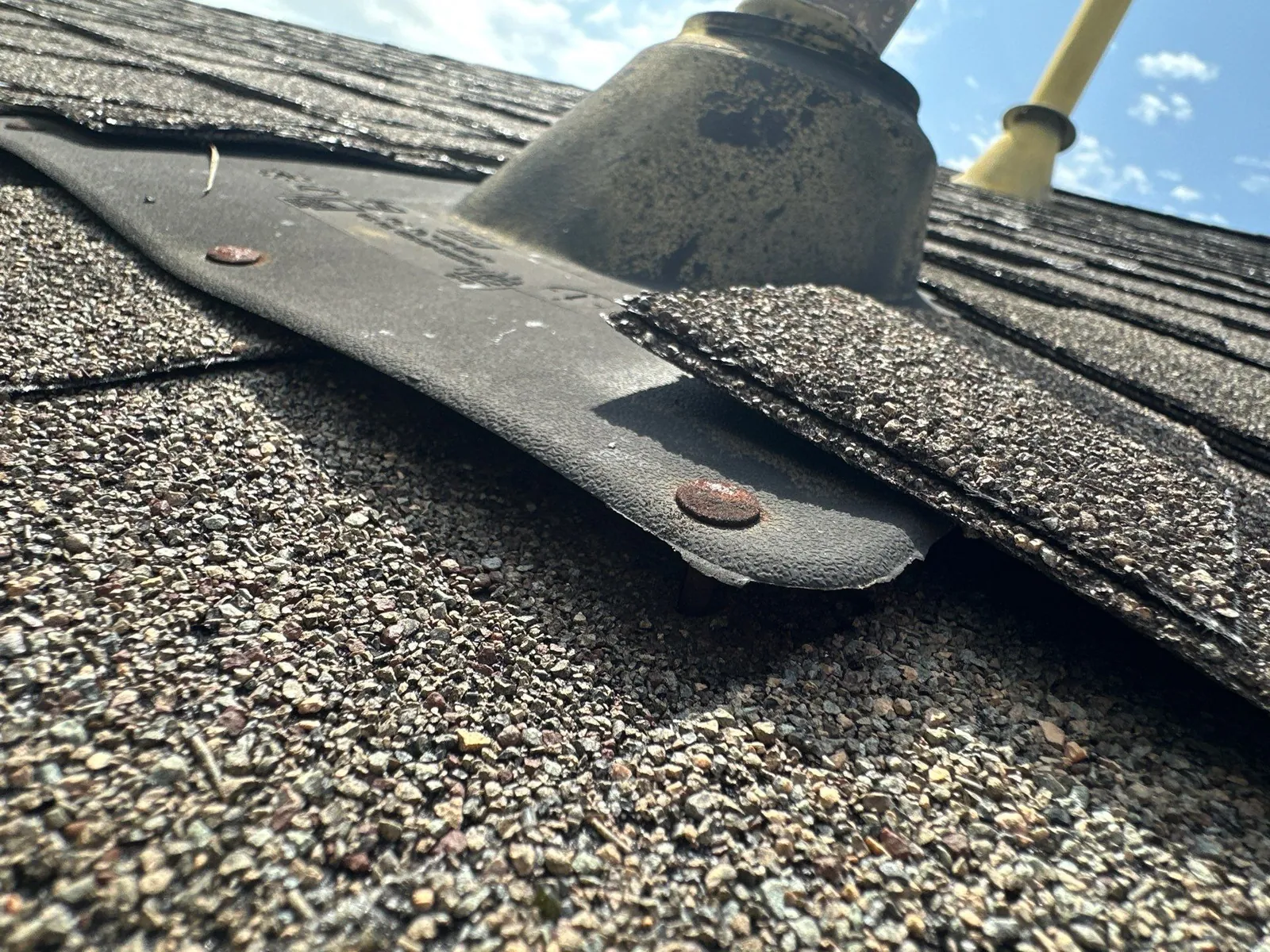 Close-up of a pipe boot flange on a shingle roof, lifting off the shingles where its rusted nail has backed out