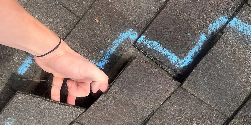 A hand lifting a shingle inside a test area marked in blue chalk on a dark shingle roof